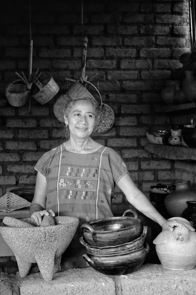 Charming black and white portrait of a Mexican woman in traditional dress surrounded by clay pots in her kitchen.